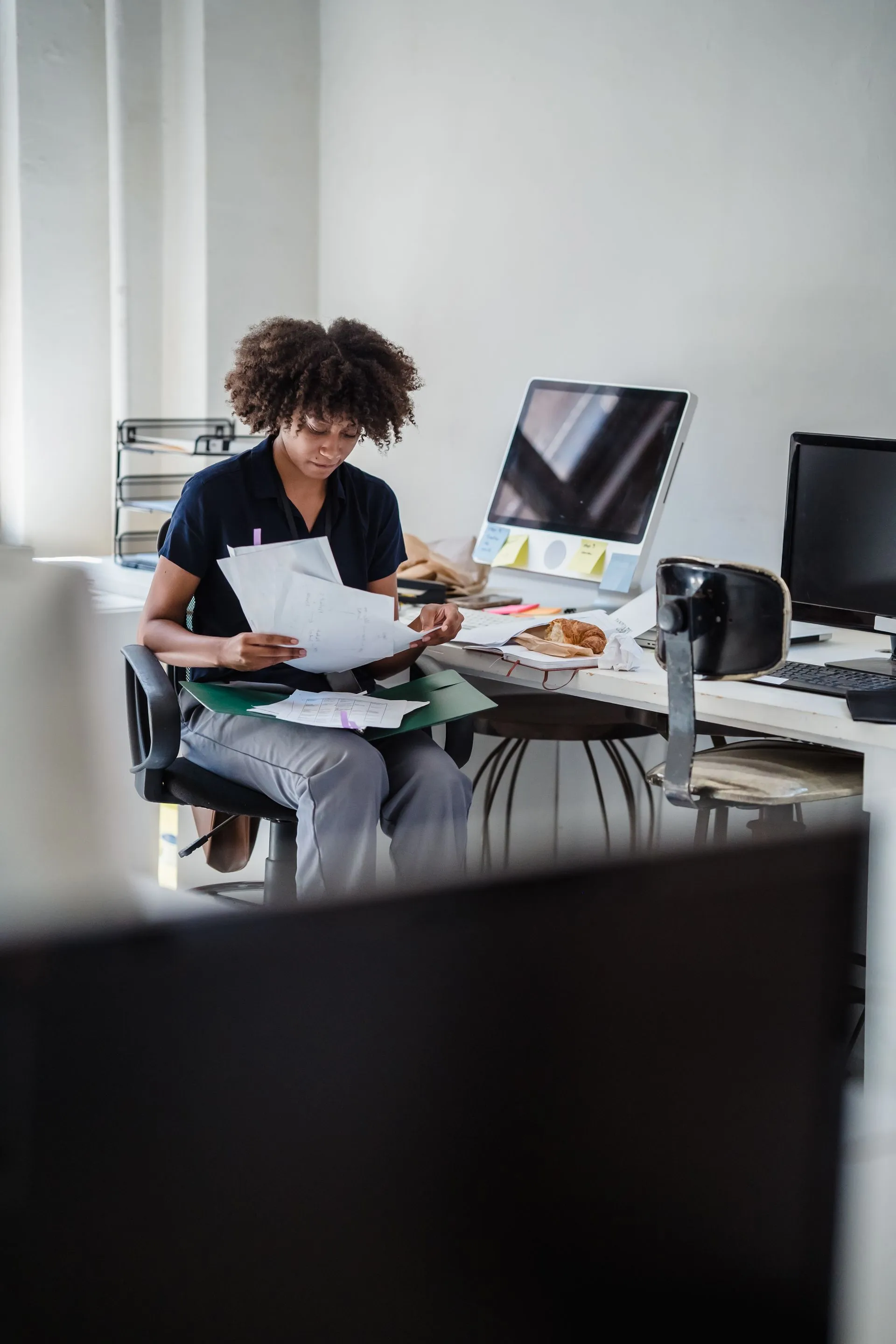 A woman reviewing paperwork at a desk with multiple monitors in an office